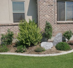 A small flower bed with some bushes on the side of a brick home